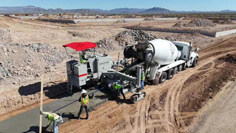 Aerial view of a Power Curber machine placing a wide concrete slab in the desert as a mixer truck feeds material and workers finish the surface with mountains in the background.