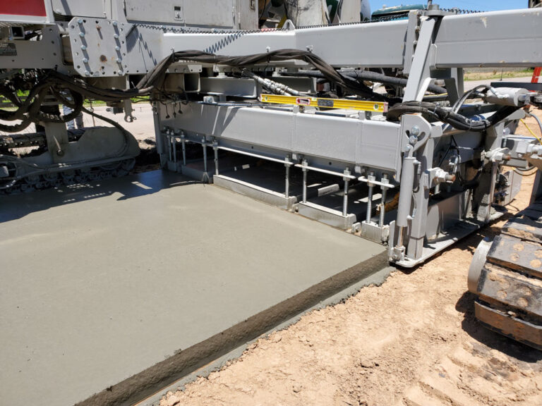 Close-up of the mold and finishing pan on a slipform machine placing a wide concrete slab.