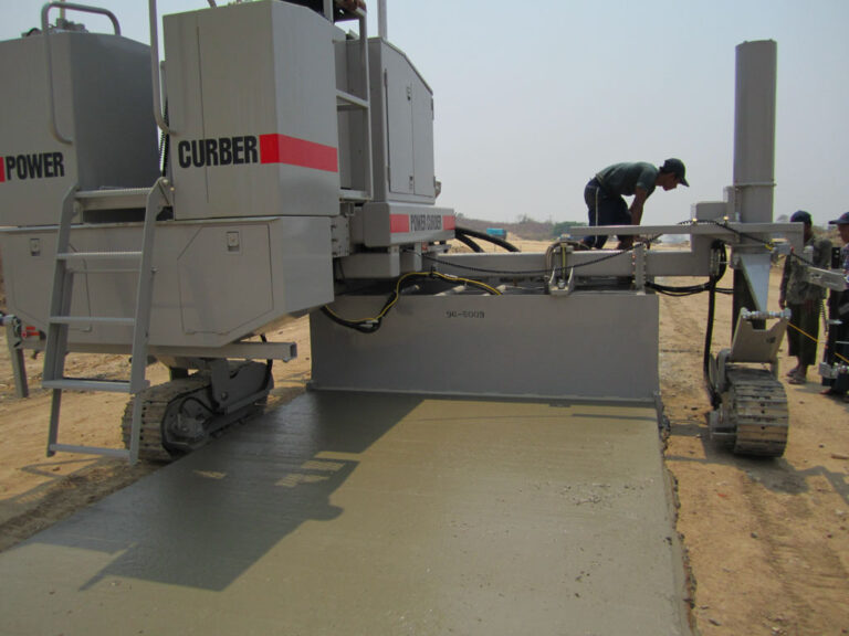 Power Curber machine placing a wide concrete slab as a worker inspects the mold and others look on.