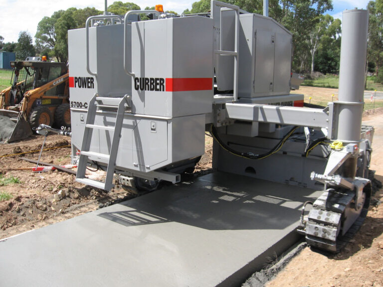 Power Curber 5700-C machine placing a wide concrete slab as a skid steer works nearby under trees.
