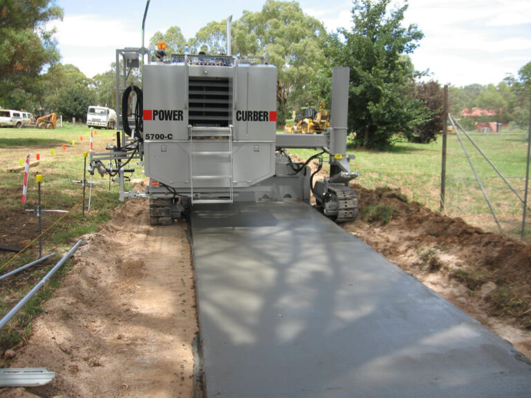 Power Curber 5700-C machine placing a concrete pathway through a grassy, tree-lined property.