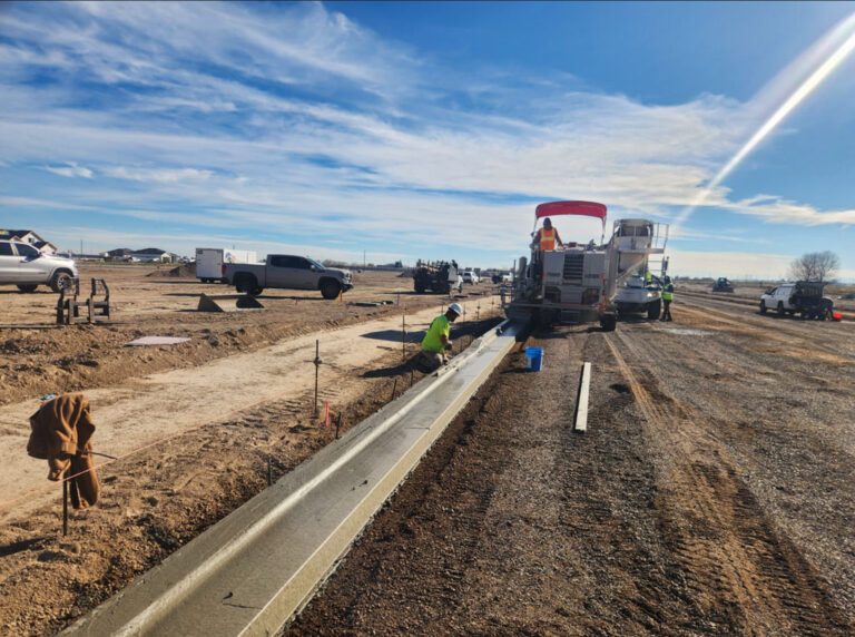 Power Curber machine with a red canopy placing a concrete curb and gutter on a desert lot as a worker finishes the surface.