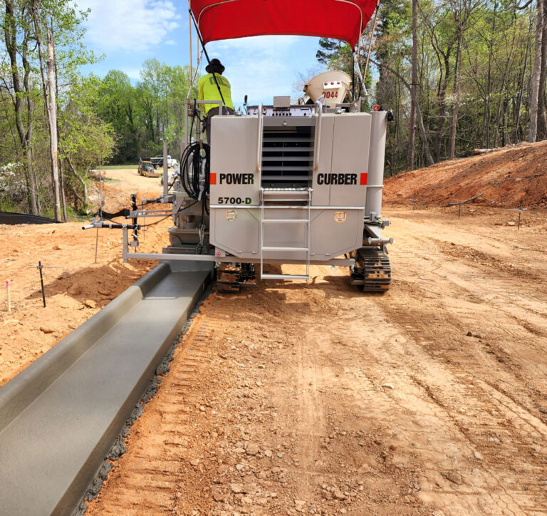 Power Curber 5700-D machine placing concrete curb and gutter along a wooded dirt road with a worker on the platform.