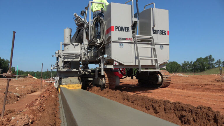 Power Curber 5700-D MAX machine placing concrete curb and gutter on a red-dirt site with a worker on the platform.