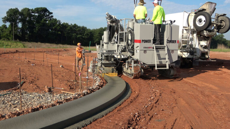 Power Curber 5700-D MAX machine placing a curved concrete curb and gutter with two workers on the platform beside a ready-mix truck.