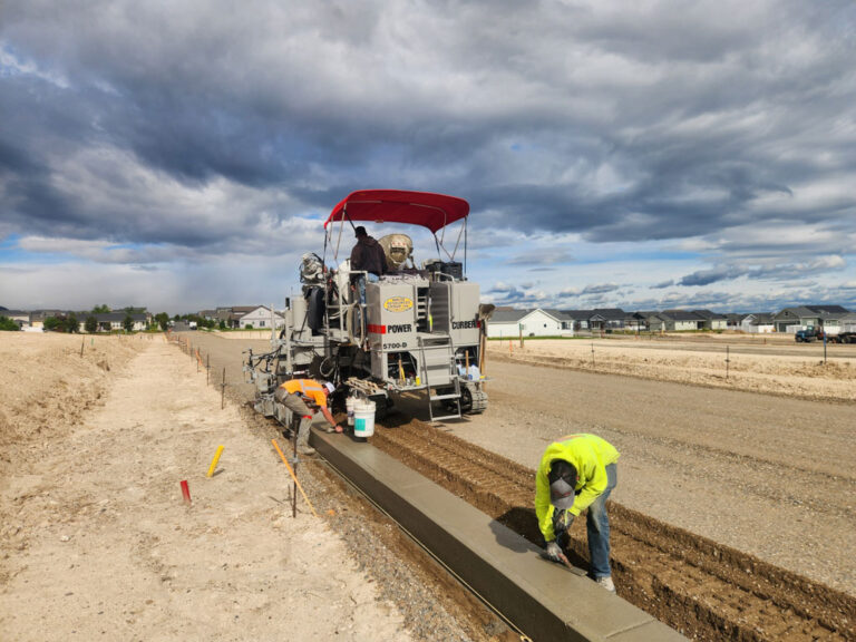 Power Curber 5700-D machine placing concrete curb and gutter in a new residential development under a cloudy sky, with two workers on site.