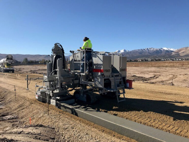 Power Curber 5700-D machine placing concrete curb and gutter with snow-capped mountains in the background and a worker on the platform.