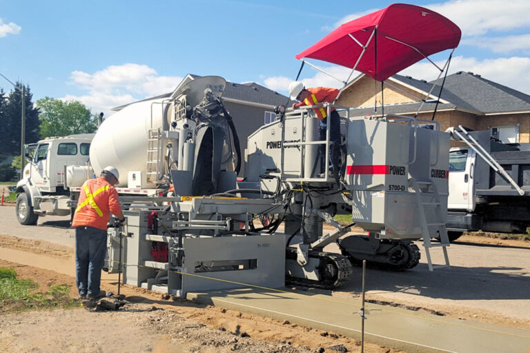 Power Curber 5700-D machine placing a monolithic curb and sidewalk as two workers guide the pour beside a mixer truck.
