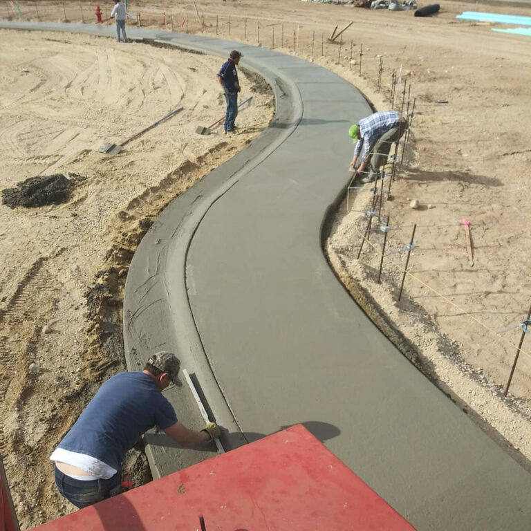 Aerial view of workers finishing a curved monolithic concrete sidewalk and curb on a sandy lot.
