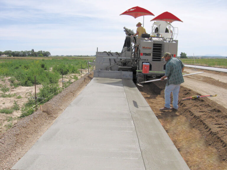Power Curber 5700-C machine branded McCoy placing a curved monolithic curb and sidewalk beside farmland, with two workers under red umbrellas.
