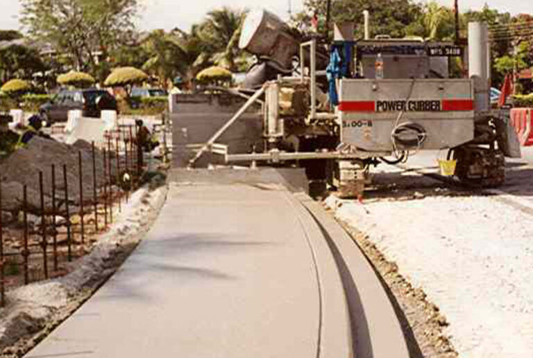 Power Curber machine placing a curved concrete sidewalk in a tropical landscaped area with palm trees.