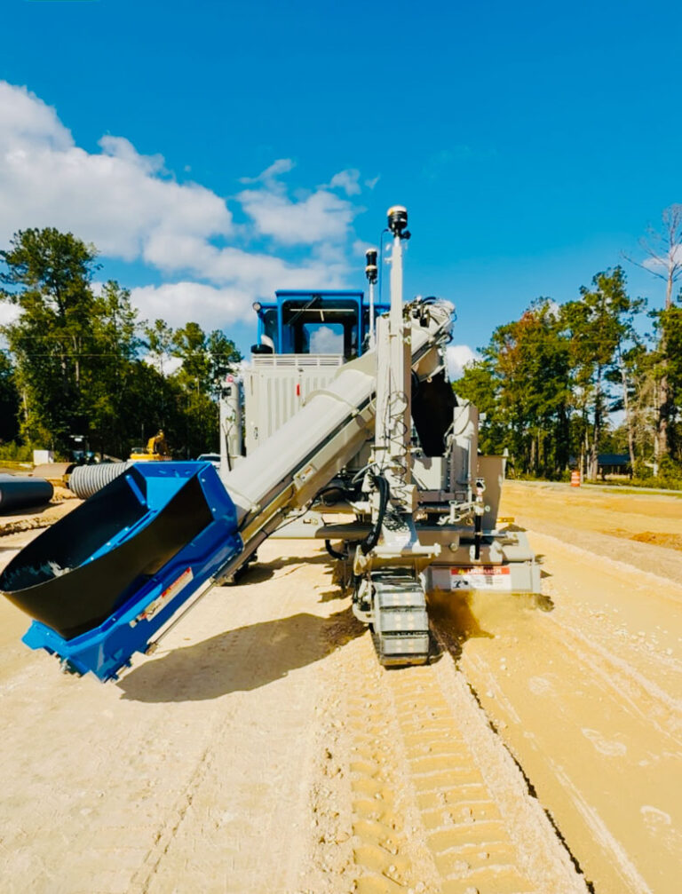 Blue-painted Power Curber machine with a curved conveyor arm parked on a dirt road, with trees in the background.