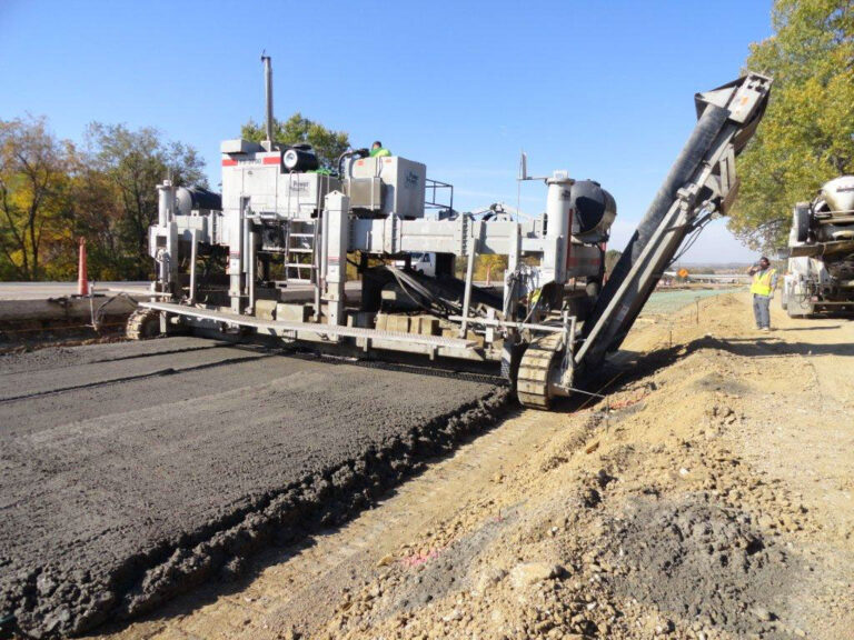 Power Paver placer/spreader machine spreading concrete on a highway lane as a ready-mix truck feeds material and a worker looks on.