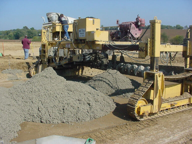 Yellow Curbmaster placer/spreader machine spreading a mound of concrete in a field as two workers monitor the process.