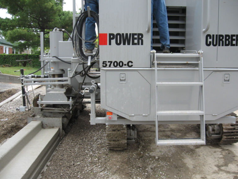 Close-up of a Power Curber 5700-C machine's mold and hydraulics beside a freshly placed concrete curb on a residential street.