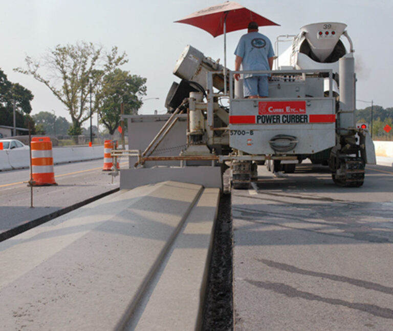 Power Curber 5700-B machine branded for Curbs Etc. placing a highway barrier wall under a red umbrella as a worker monitors the equipment.