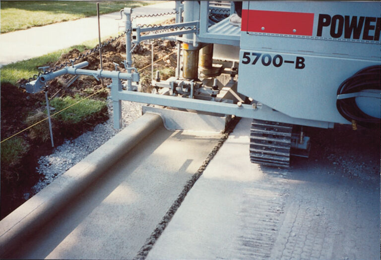 Close-up of a Power Curber 5700-B machine's mold forming a curved concrete curb and sidewalk.