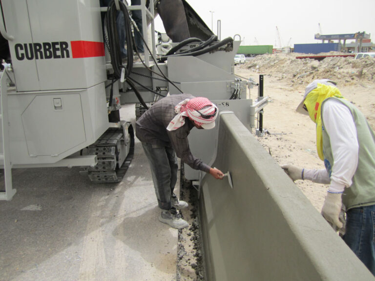 Power Curber machine forming a concrete barrier wall as two workers in headscarves and safety gear finish the surface at an overseas job site.
