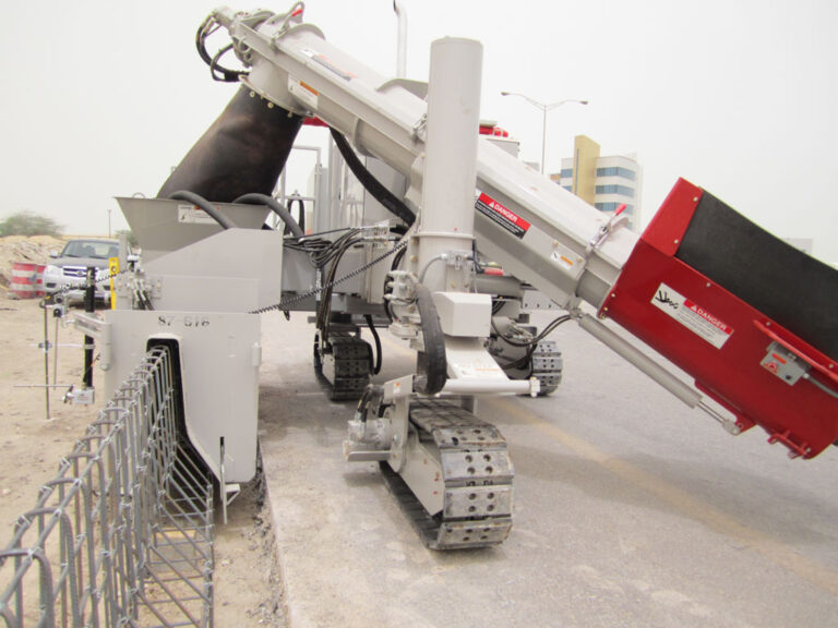 Close-up of a Power Curber machine's conveyor arm and red hopper feeding concrete beside a chain-link fence.