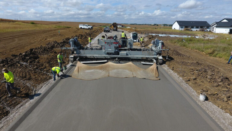 Aerial view of a Power Paver SF-2700 placing a residential street slab as workers finish the surface and a burlap curing blanket trails behind.