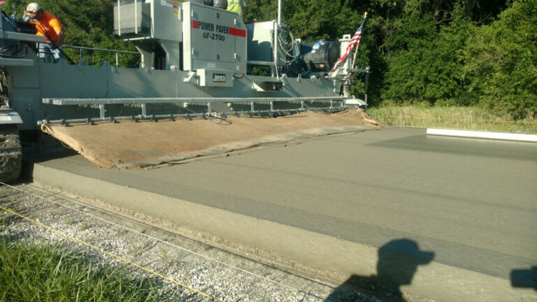 Power Paver SF-2700 branded Jones placing a concrete road slab with a burlap curing blanket trailing behind and an American flag mounted on the machine.