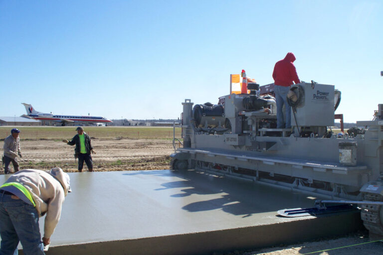 Power Pavers SF-2700 machine placing a wide concrete slab at an airport apron with a parked airplane and workers finishing the surface.