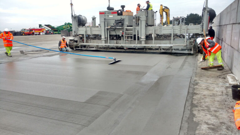 Power Pavers SF-3000 machine placing a wide concrete slab as workers finish the surface along a barrier wall.