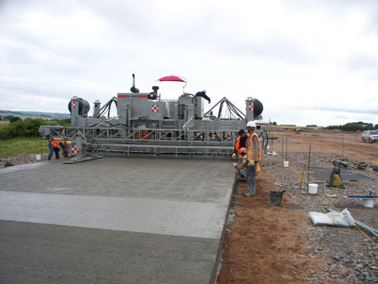 Power Pavers SF-3000 machine placing a wide concrete slab under a red umbrella as workers finish the edge on a rural site.