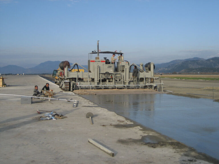 Power Pavers SF-3000 machine placing a wide concrete slab beside a canal with mountains in the background and workers finishing the surface.