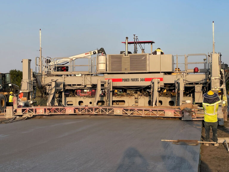 Power Pavers SF-3404 machine placing a wide concrete slab at dusk, with a worker in a high-visibility vest overseeing the pour.