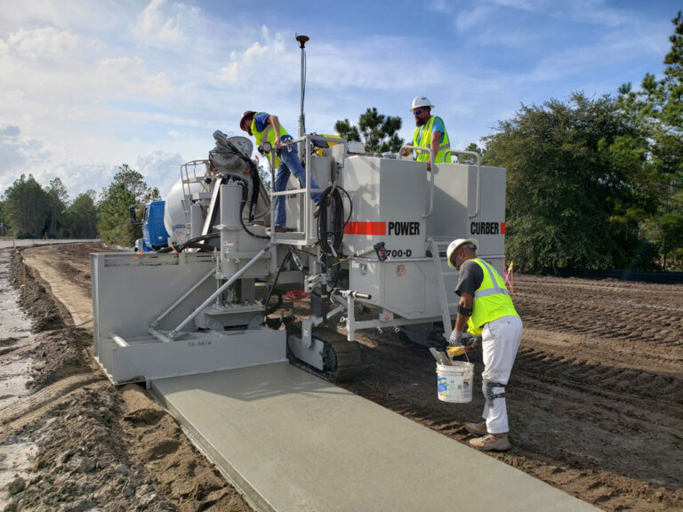 Power Curber 5700-D machine placing a concrete sidewalk with two workers on the platform and a third finishing the edge with a bucket of tools.