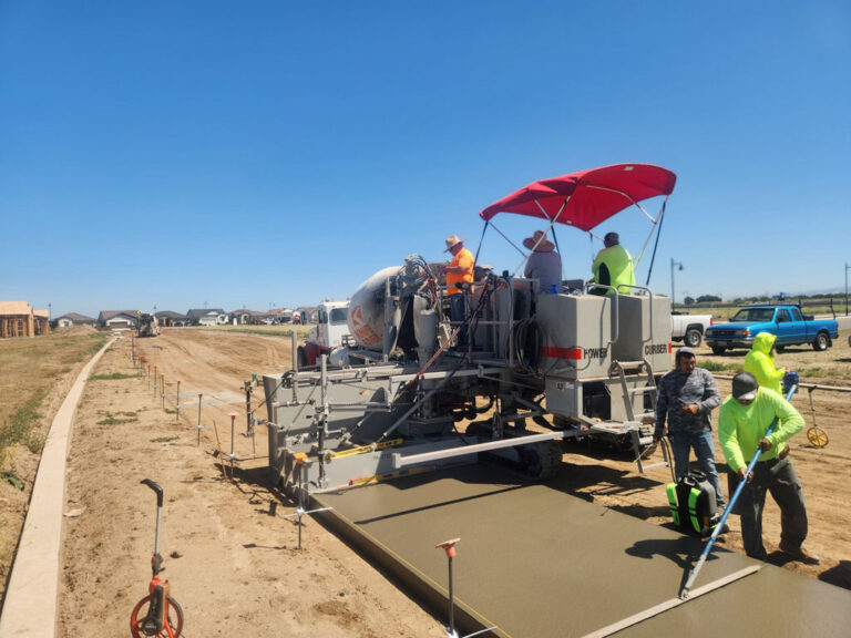 Power Curber machine placing a wide concrete sidewalk as several workers in high-visibility gear finish the surface and a mixer truck feeds material.