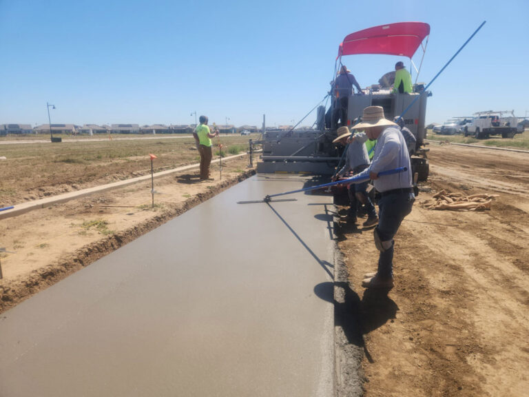 Power Curber machine placing a wide concrete sidewalk in a new subdivision as several workers in straw hats finish the surface with hand tools.