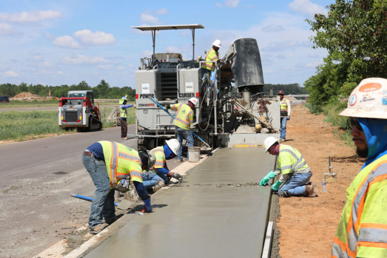 Power Curber 5704-D MAX machine placing a highway shoulder as several workers finish the concrete and place rebar.