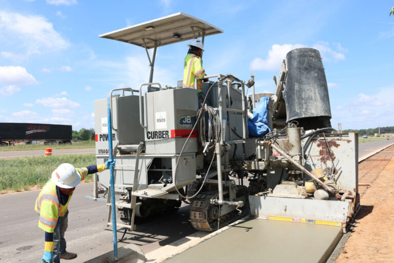 Power Curber 5704-D MAX machine branded JB Power Curber placing a concrete lane beside a highway, with a worker guiding the pour.