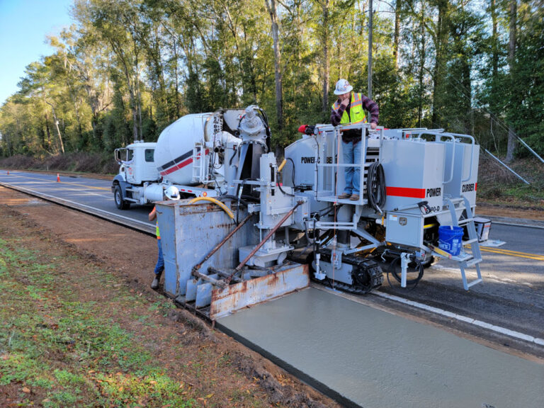 Power Curber 5700-D machine placing a rural roadside concrete path as a ready-mix truck feeds material and a worker monitors the equipment.