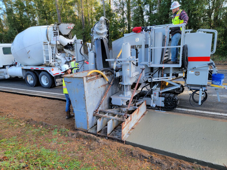 Power Curber machine placing a concrete sidewalk with an integral drain trough as a ready-mix truck feeds material and workers guide the pour.