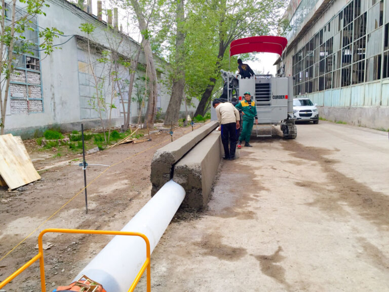 Power Curber machine forming a concrete slot drain trough beside a pipe, with two workers inspecting the finished section.