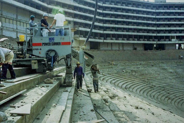 Power Curber 5700-B machine branded ICC forming curved stadium seating risers as workers guide the pour inside a large stadium under construction.