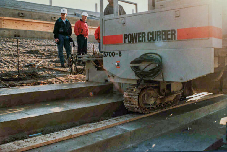 Close-up of a Power Curber 5700-B machine forming stadium seating risers as three workers observe the pour.