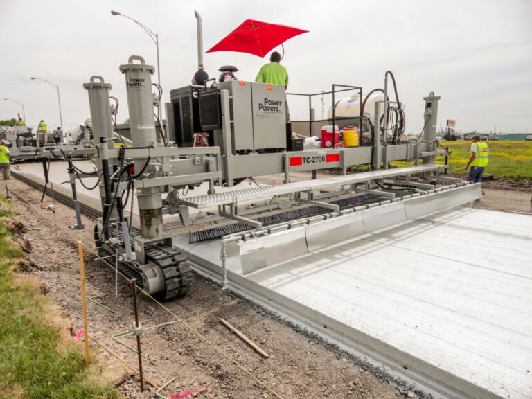 Power Pavers TC-2700 texture and curing machine finishing a wide concrete slab under a red umbrella as a worker operates the controls.