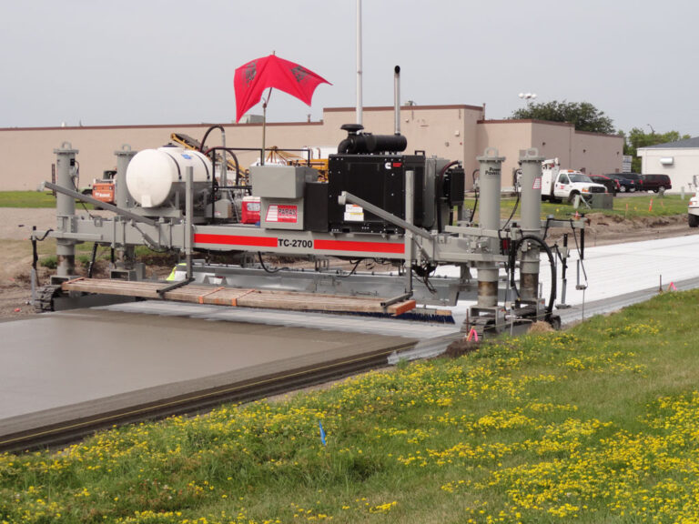 Power Pavers TC-2700 texture and curing machine applying a broom finish to a concrete slab beside a wildflower field.
