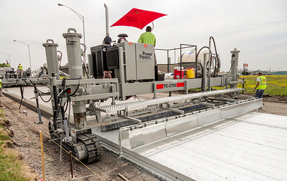 Power Pavers TC-2700 texture and curing machine finishing a concrete slab under a red umbrella with a worker nearby.