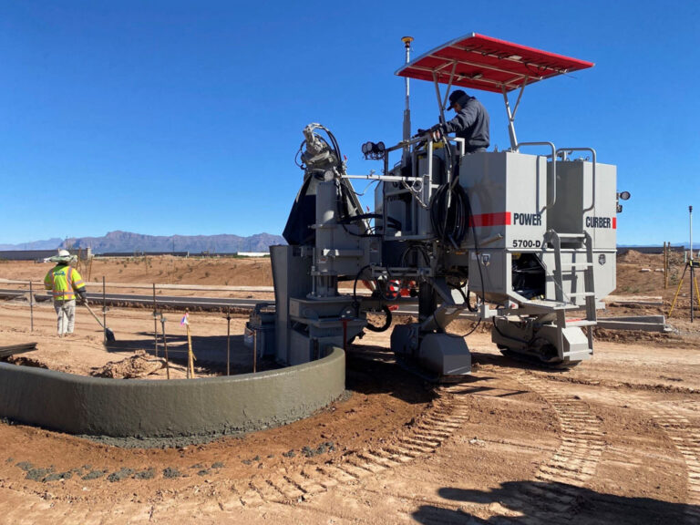 Power Curber 5700-D machine forming a tight-radius concrete curb in the desert as a worker in a high-visibility vest looks on.