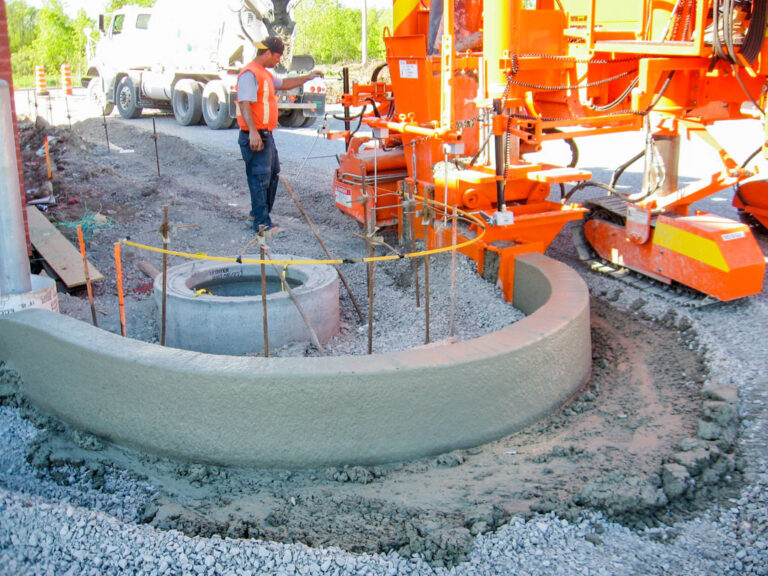 Power Curber machine forming a curved concrete barrier around a manhole as a worker in an orange vest inspects the pour beside a mixer truck.
