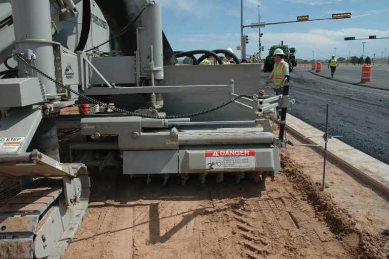 Close-up of a Power Curber trimmer attachment with a warning label, positioned beside a roadway with traffic signals in the background.